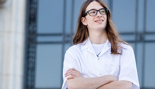 A high school senior with long brown hair and glasses stands confidently with arms crossed, wearing a white lab coat. They are outdoors in front of a building with tall columns and large windows, reminiscent of classic senior pictures in Detroit.