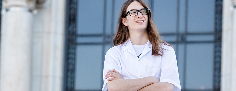 A high school senior with long brown hair and glasses stands confidently with arms crossed, wearing a white lab coat. They are outdoors in front of a building with tall columns and large windows, reminiscent of classic senior pictures in Detroit.