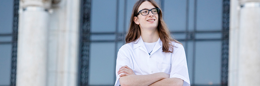 A high school senior with long brown hair and glasses stands confidently with arms crossed, wearing a white lab coat. They are outdoors in front of a building with tall columns and large windows, reminiscent of classic senior pictures in Detroit.