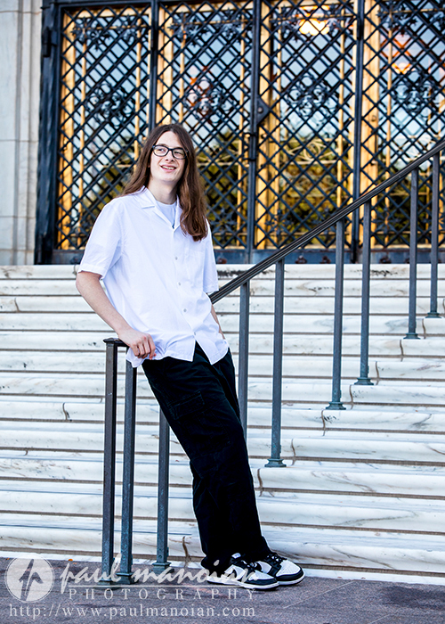 A high school senior boy with long hair and glasses leans casually against a staircase railing outside a building with ornate, metal lattice doors, captured for senior pictures in Detroit. They wear a white short-sleeved button-up shirt, dark pants, and sneakers. A photography watermark is in the lower left corner of the image.