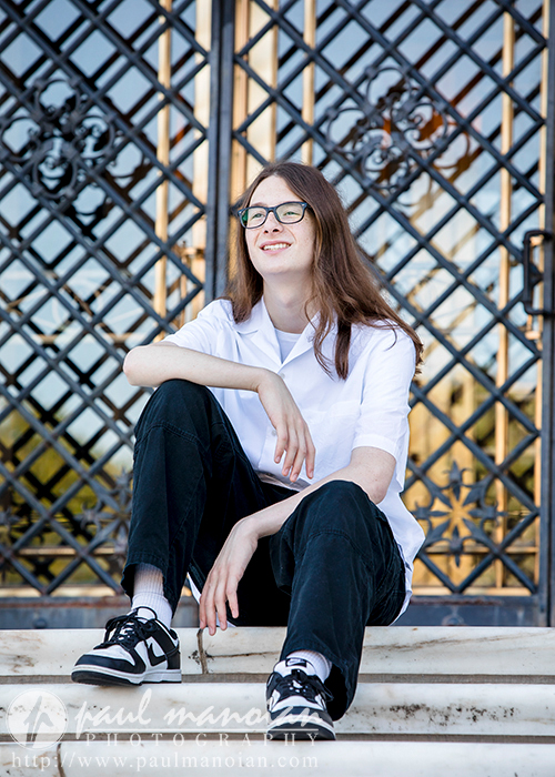 A high school senior with long hair and glasses sits on outdoor steps in front of a decorative metal gate, perfect for senior pictures in Detroit. They wear a white shirt, black pants, and black-and-white sneakers. They have a relaxed expression and are looking slightly to the side.