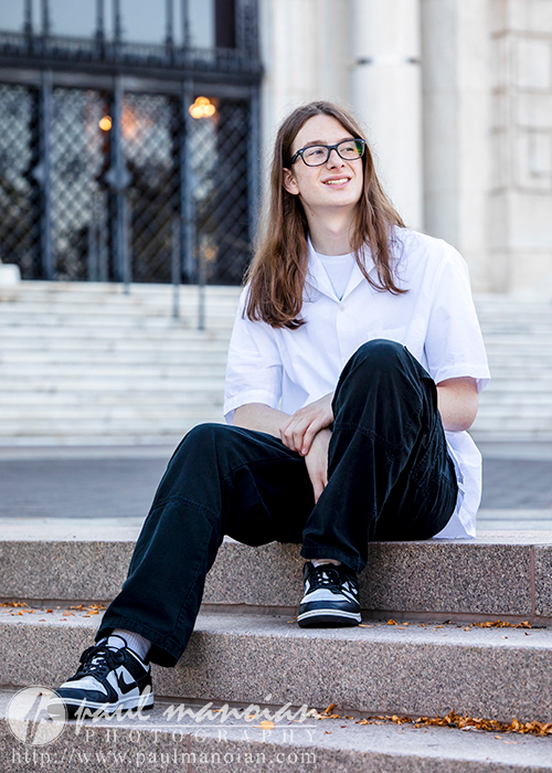 A high school senior with long hair and glasses sits on outdoor steps, smiling while gazing to the side. They wear a white shirt, black pants, and sneakers. The background features a large building with ornate doors. Fallen leaves dot the steps around them, adding charm to these senior pictures in Detroit.