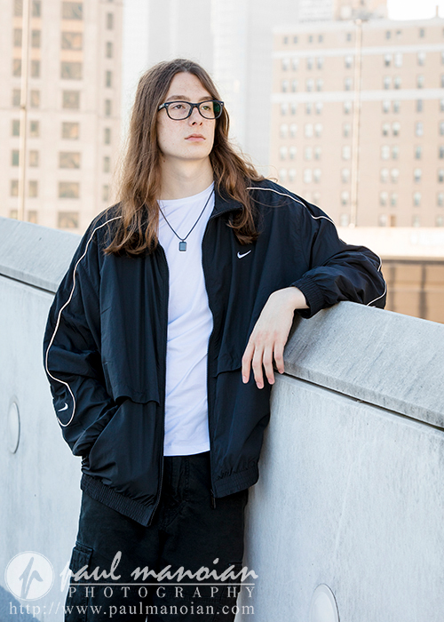 A high school senior with long hair and glasses is leaning against a concrete ledge in an urban setting, perfect for senior pictures in Detroit. They are wearing a black jacket, white t-shirt, and dark pants. Buildings are visible in the background. The image features a logo and URL for Paul Manoian Photography.