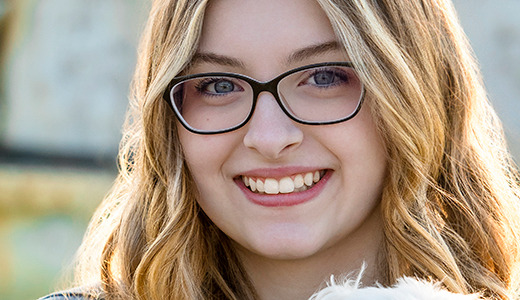 A high school senior girl with long, wavy blonde hair and black-framed glasses smiles at the camera, holding a small white dog. The background, slightly blurred but featuring a stone structure, gives the photograph an elegant touch reminiscent of Trenton senior pictures.