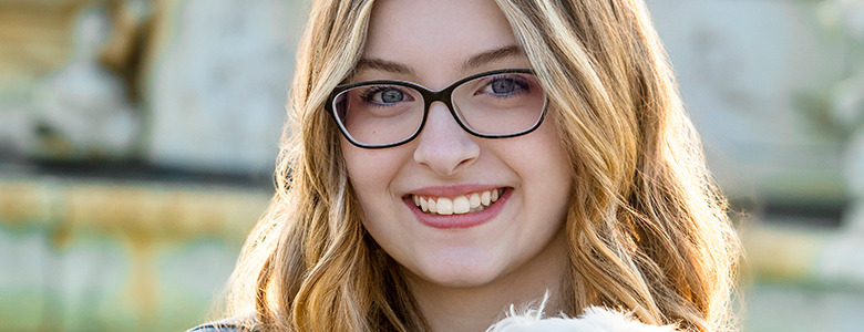 A high school senior girl with long, wavy blonde hair and black-framed glasses smiles at the camera, holding a small white dog. The background, slightly blurred but featuring a stone structure, gives the photograph an elegant touch reminiscent of Trenton senior pictures.