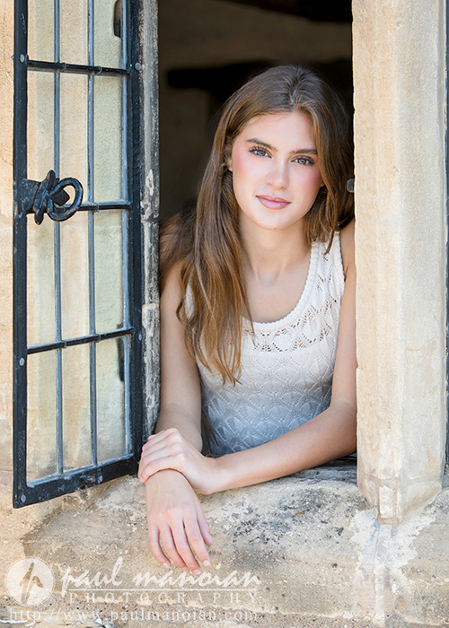A high school senior with long, brown hair and a white sleeveless top leans on a stone windowsill, framed by an open window with a decorative metal grill. She gazes directly at the camera with a gentle expression, capturing a perfect moment in her Trenton senior portraits.