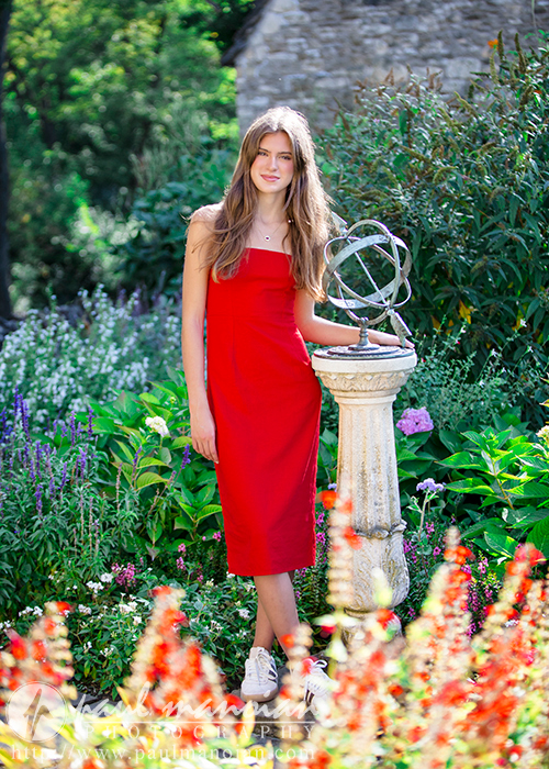A high school senior in a red dress stands beside a stone pedestal with a spherical metal sculpture in a lush garden, perfect for Trenton senior portraits. She has long brown hair and smiles softly amid the vibrant green plants and colorful flowers, with a stone building visible in the background.