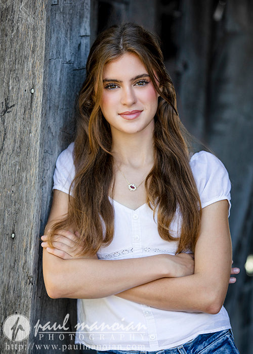 A high school senior with long brown hair stands against a wooden wall with her arms crossed. She wears a white blouse and a simple necklace. Smiling softly at the camera, her image exudes confidence, captured perfectly in these Trenton senior portraits. A professional watermark adorns the lower left corner.