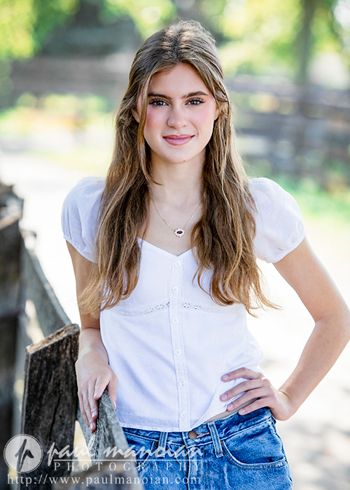A high school senior with long, wavy brown hair smiles softly while standing outdoors. She wears a white, short-sleeve blouse and blue jeans, resting one hand on a wooden fence. The background is blurred, highlighting greenery and a wooden structure in a bright setting—perfect for Trenton senior portraits.