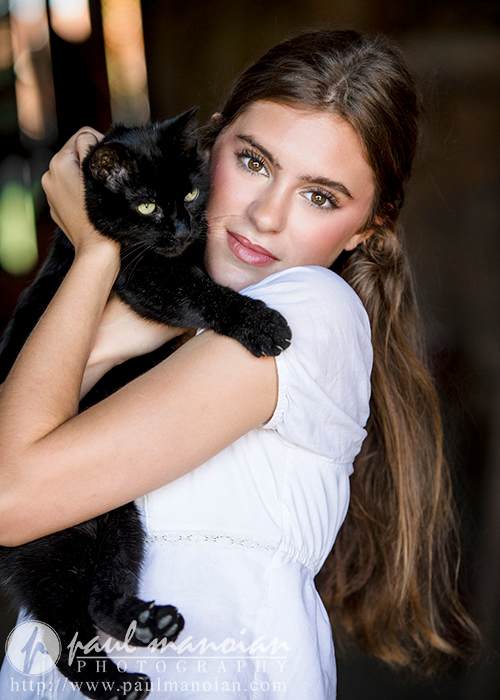 A high school senior in a white dress lovingly holds a black cat close to her face. She gazes directly at the camera with a serene expression, perfect for Trenton senior portraits. The cat, with piercing green eyes, looks slightly to the side. The background is blurred, highlighting the subjects.