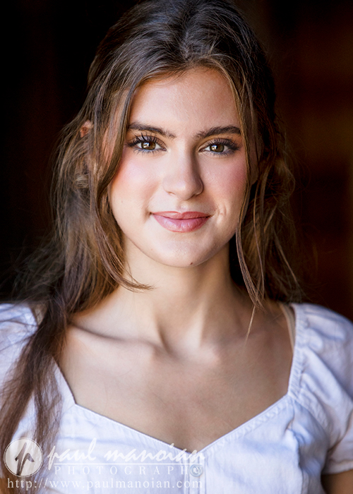 Portrait of a high school senior with long brown hair, wearing a white blouse, smiling softly at the camera. The image has a slightly dark background, emphasizing her face. The bottom of the photo includes text and a logo from Trenton Senior Portraits photography studio.