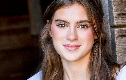 A high school senior girl with long brown hair and a warm smile stands against a rustic wooden backdrop, perfect for Trenton senior portraits. She has light makeup with rosy cheeks, and her eyes are bright and expressive. She's wearing a white top, and the natural light highlights her features beautifully.