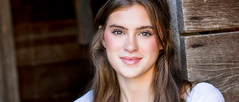 A high school senior girl with long brown hair and a warm smile stands against a rustic wooden backdrop, perfect for Trenton senior portraits. She has light makeup with rosy cheeks, and her eyes are bright and expressive. She's wearing a white top, and the natural light highlights her features beautifully.
