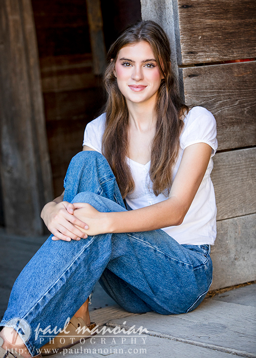 High school senior girl with long brown hair sits on wooden steps, smiling. She's wearing a white short-sleeve top and blue jeans. The background features a wooden wall. Text at the bottom reads "Paul Manoian Photography" and displays a URL, highlighting Trenton senior portraits.