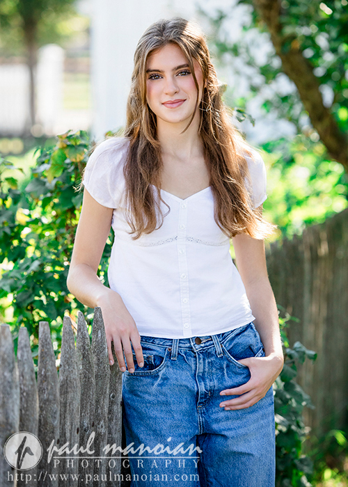 A high school senior girl with long, light brown hair, wearing a white short-sleeve blouse and blue jeans, leans against a wooden fence outdoors. She smiles softly, with greenery and sunlight in the background. The text "Paul Manoian Photography - Trenton senior portraits" and website link are visible at the bottom.