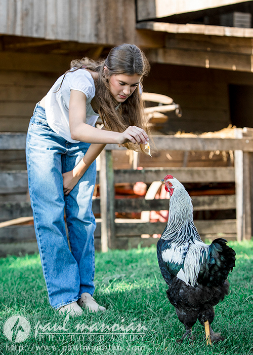 A high school senior with long hair is bending over to feed a black-and-white rooster outside. She is wearing a white t-shirt and blue jeans, looking perfectly candid for Trenton senior portraits. In the background, there's a wooden structure and some greenery.