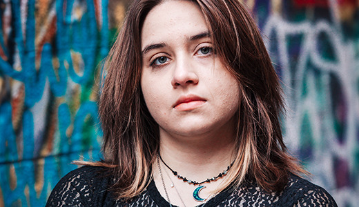 A high school senior with shoulder-length hair stands in front of a colorful graffiti wall, capturing the vibe of Ann Arbor senior pictures. They are wearing a black lace top and layered necklaces, including one with a crescent moon pendant, looking directly at the camera with a neutral expression.