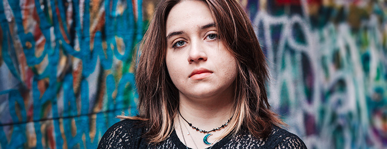 A high school senior with shoulder-length hair stands in front of a colorful graffiti wall, capturing the vibe of Ann Arbor senior pictures. They are wearing a black lace top and layered necklaces, including one with a crescent moon pendant, looking directly at the camera with a neutral expression.