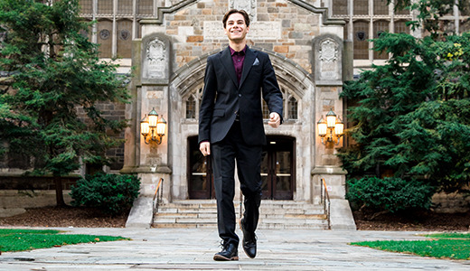 A high school senior boy in a black suit walks confidently on a cobblestone path toward a historic building with an arched entrance and ornate stonework, framed by lush greenery. The scene, perfect for Ann Arbor senior portraits, conveys a sense of achievement and elegance.