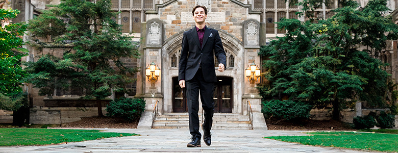 A high school senior boy in a black suit walks confidently on a cobblestone path toward a historic building with an arched entrance and ornate stonework, framed by lush greenery. The scene, perfect for Ann Arbor senior portraits, conveys a sense of achievement and elegance.