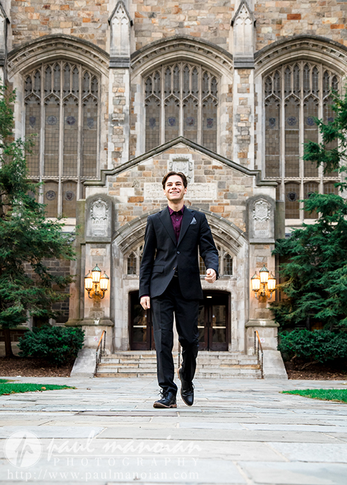 A high school senior boy in a dark suit walks confidently on a stone path lined with greenery, reminiscent of an Ann Arbor senior portrait, with a historic building featuring arched windows and a wooden door in the background.