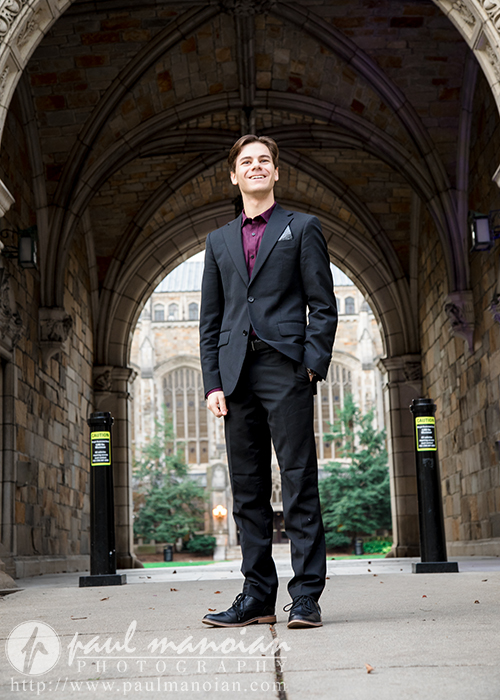 A high school senior boy in a black suit stands smiling under a stone archway with gothic architectural features, capturing the essence of Ann Arbor senior portraits. Trees and a large building with arched windows are visible in the background, creating a timeless scene.