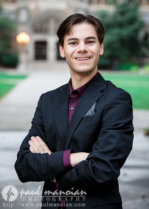 A high school senior boy in a black suit and purple shirt stands outdoors with arms crossed, smiling confidently against the backdrop of a blurred building and greenery—capturing the essence of Ann Arbor senior portraits.