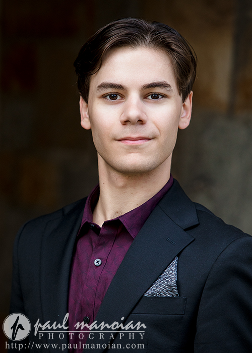 A high school senior boy with short, dark hair wearing a black suit jacket over a dark maroon shirt stands facing the camera. The background is softly blurred, giving the image the polished feel of Ann Arbor senior portraits.