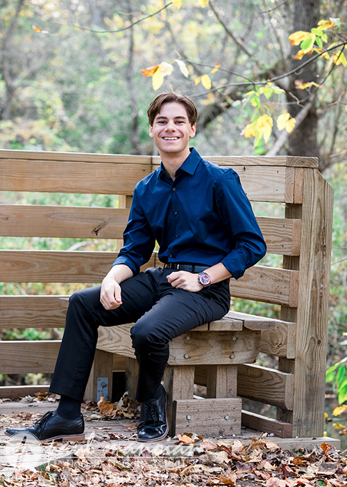 A high school senior wearing a blue shirt and black pants sits on a wooden bench in a park, perfect for capturing Ann Arbor senior portraits. The ground is adorned with fallen leaves, and trees with vibrant autumn foliage provide a stunning backdrop.