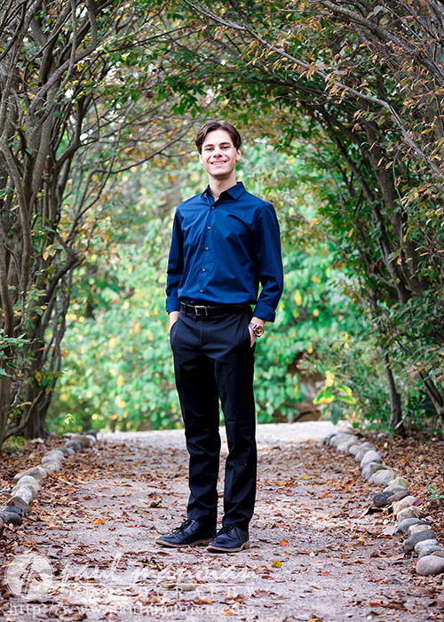 A high school senior in a blue shirt and black pants stands on a cobblestone pathway surrounded by trees and greenery, smiling. Perfect for Ann Arbor senior portraits, the trees form a natural arch overhead.