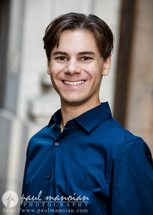 A high school senior with short, dark hair is smiling and wearing a blue shirt, perfectly capturing the essence of Ann Arbor senior portraits. The softly blurred background focuses attention on the individual. The image features a watermark from Paul Manoian Photography at the bottom.