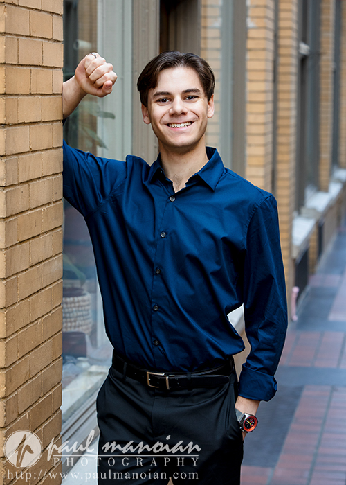 A smiling high school senior boy in a navy blue shirt and black pants leans against a brick wall, capturing the essence of Ann Arbor senior portraits. With short brown hair and a wristwatch, he stands on reddish-brown and black tiles that add charm to this alleyway moment.