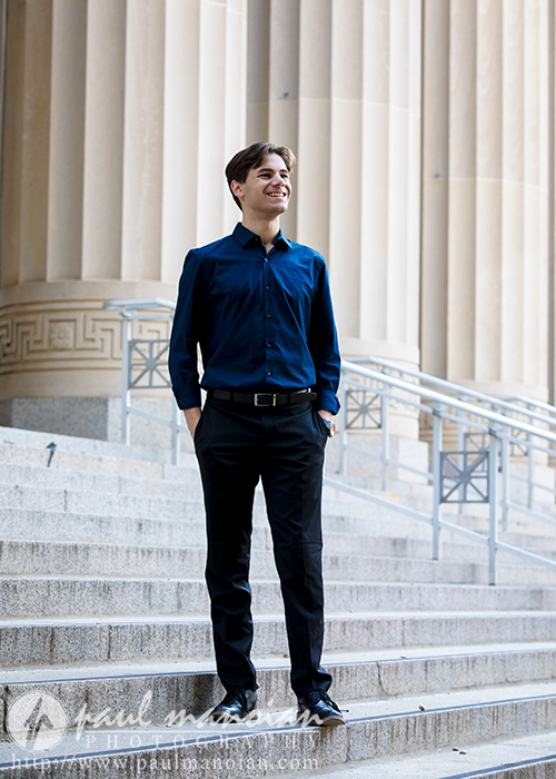 A high school senior in a blue shirt and black pants stands on a set of steps in front of a building with tall columns, reminiscent of classic Ann Arbor architecture. Smiling and looking into the distance, they capture the essence of senior portraits with confidence and grace.