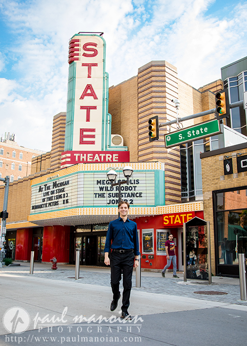 A high school senior strolls along the sidewalk in front of the State Theatre, where the marquee announces upcoming shows. The theatre's retro signage, a favorite backdrop for Ann Arbor senior portraits, and surrounding buildings stand out under a clear blue sky.
