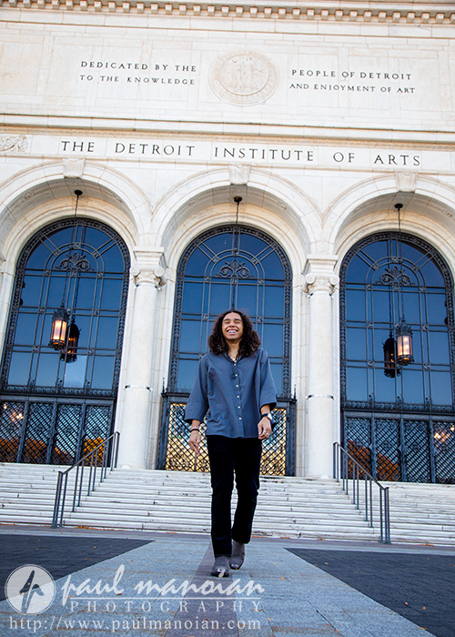 A high school senior with long hair and a blue shirt stands smiling, skateboard in hand, in front of the Detroit Institute of Arts. The steps lead up to large arched windows and a stone facade with an inscription at the top, creating the perfect backdrop for senior pictures.