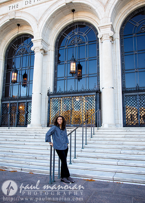 A high school senior stands on the steps in front of a grand building with tall arched windows and ornate iron grilles, casually holding a skateboard. The entrance, decorated with hanging lanterns, creates an ideal backdrop for senior pictures as the scene is bathed in natural light.