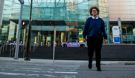 A high school senior with curly hair, wearing a blue sweater and black pants, walks across a crosswalk towards a building with a modern glass facade. There are scooters lined up on the right side of the image.