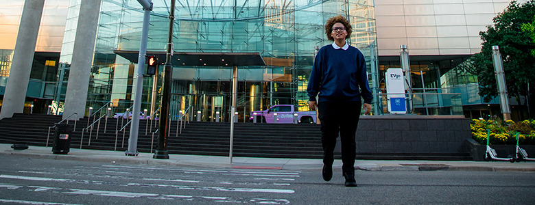 A high school senior with curly hair, wearing a blue sweater and black pants, walks across a crosswalk towards a building with a modern glass facade. There are scooters lined up on the right side of the image.