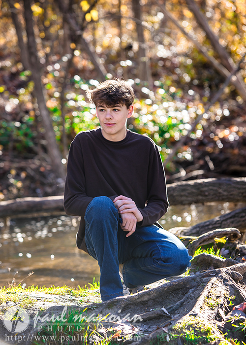 A high school senior with short hair kneels on a log by a stream, surrounded by the sunlit splendor of a fall forest. They wear a dark sweater and blue jeans, capturing an introspective moment perfect for senior pictures.