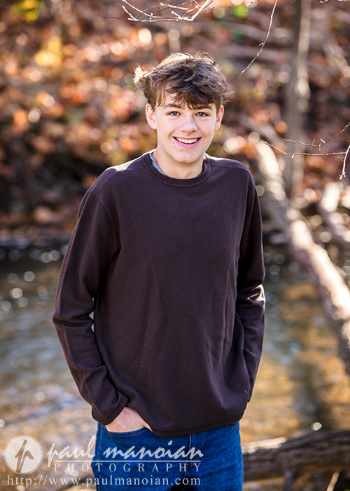 A high school senior with short brown hair smiles near a stream, capturing the essence of fall senior pictures. They're clad in a brown long-sleeve shirt and jeans, framed by blurred autumn leaves and bare branches in the background, perfectly embodying this memorable moment.
