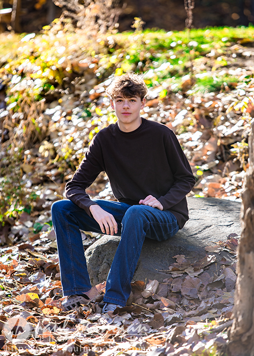 A high school senior with short brown hair sits on a rock in a forested area, capturing their serene essence in fall senior pictures. Wearing a dark sweater and blue jeans, they are bathed in sunlight filtering through the trees, which illuminates the fallen autumn leaves around them.