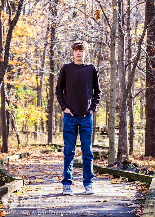 A high school senior wearing a dark sweater and jeans stands on a wooden path in a forest during autumn, perfect for capturing fall senior pictures. The ground and trees are covered with fallen leaves, creating an idyllic backdrop for this memorable moment.