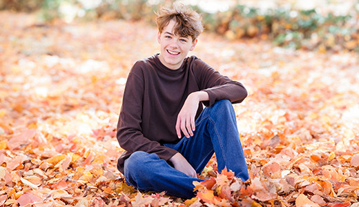 A high school senior sitting in a field of colorful autumn leaves, smiling at the camera, captures the essence of fall senior pictures. They are wearing a dark long-sleeve shirt and blue jeans. The background features blurred trees, creating a serene fall atmosphere.