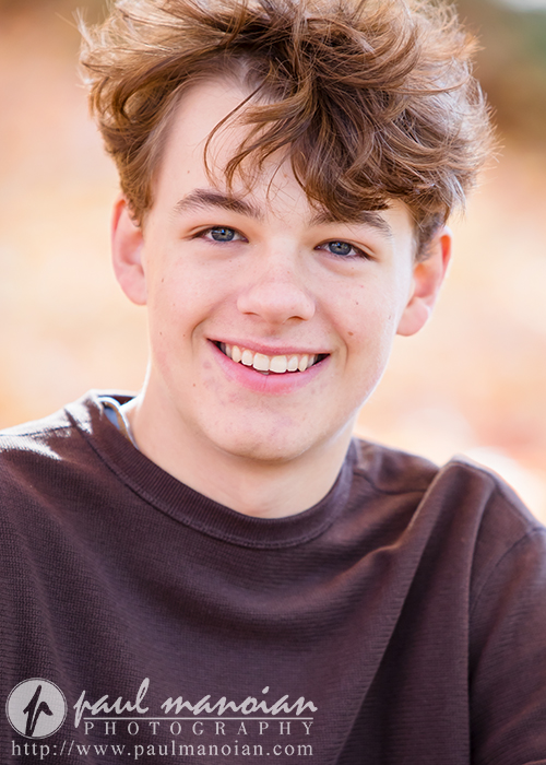 A smiling high school senior with wavy brown hair dons a brown shirt, capturing their fall senior picture against a blurred autumnal backdrop. Photography credit is subtly visible at the bottom of the image.