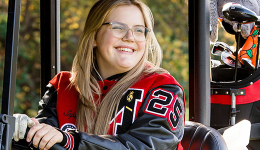 A high school senior with long blonde hair and glasses smiles while sitting in a golf cart, capturing her Golf Senior Pictures. She's wearing a red and black varsity jacket with the number 20 visible. Golf clubs are in the back of the cart, surrounded by Ann Arbor's stunning autumn foliage.