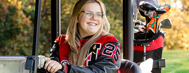 A high school senior with long blonde hair and glasses smiles while sitting in a golf cart, capturing her Golf Senior Pictures. She's wearing a red and black varsity jacket with the number 20 visible. Golf clubs are in the back of the cart, surrounded by Ann Arbor's stunning autumn foliage.