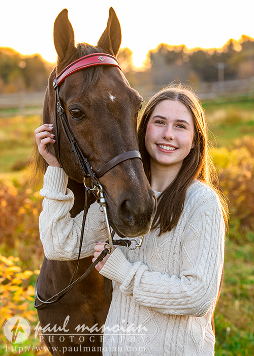 A high school senior in a white sweater smiles while standing beside a brown horse in a field during sunset, capturing the essence of autumn. With gentle hands on the bridle, this picturesque moment is perfect for senior pictures, blending warmth and nature's beauty through photography.