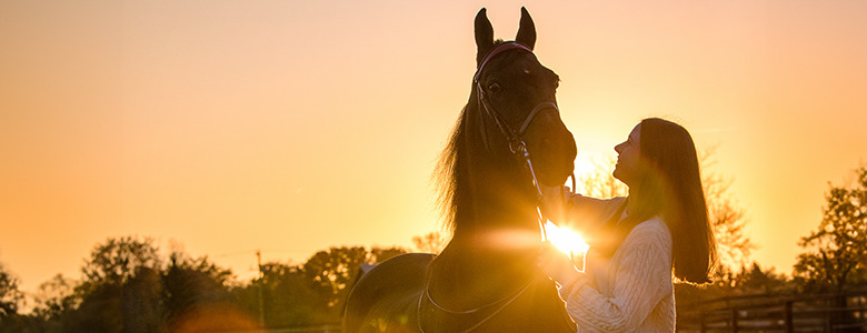 A high school senior stands beside a horse at sunset, smiling as she gently holds its bridle. The scene, reminiscent of timeless senior pictures, captures the sun's warm glow creating a serene and peaceful setting with silhouetted trees in the background.