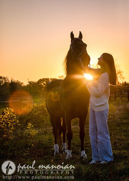 A woman poses with a horse in a field at sunset, perfect for senior pictures. The sun creates a warm glow, highlighting the silhouette and casting soft light on her sweater and flared jeans—an exquisite moment captured through photography.