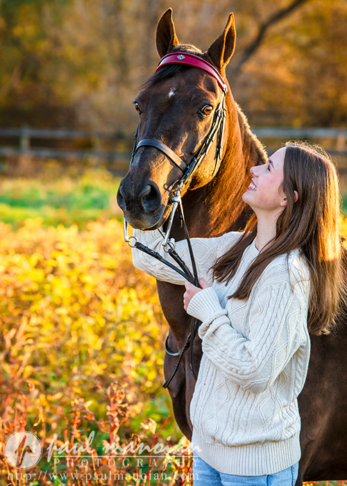 In a stunning example of equine photography, a high school senior with long hair smiles beside a dark horse, holding its reins. They stand outdoors in a sunlit autumn setting with golden foliage, perfect for capturing unforgettable senior pictures. She wears a cream sweater, blending beautifully with the scene.